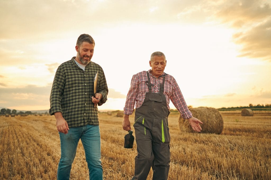 Proud mid-adult hardworking farmer standing in the wheat field on his organic farm with tablet in his hand and having a discussion about Borrowing Against Life Insurance with a senior farmer