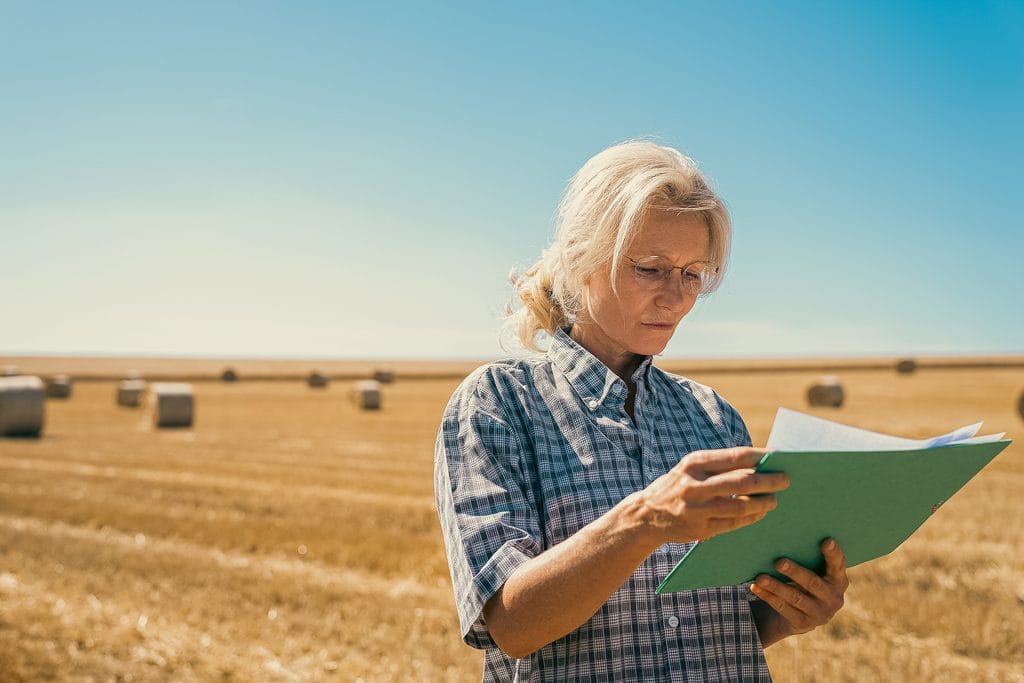 Mid-shot of woman farmer standing in agricultural field reading technical report after harvest
